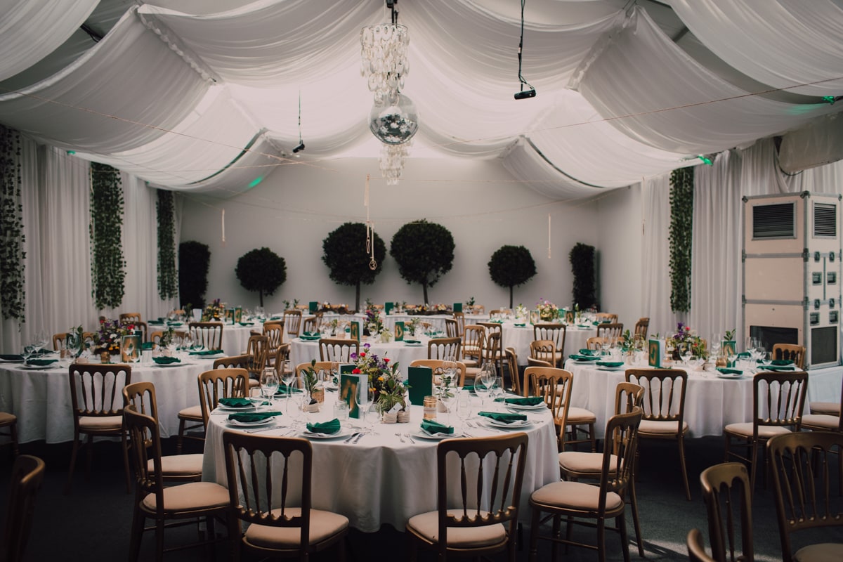 © Adrian Almasan | www.adrianalmasan.com Elegantes Festzelt mit runden Tischen, dekoriert mit grünen Servietten und Blumenarrangements, umgeben von Pflanzen und einem stilvollen, drapierten Dach.