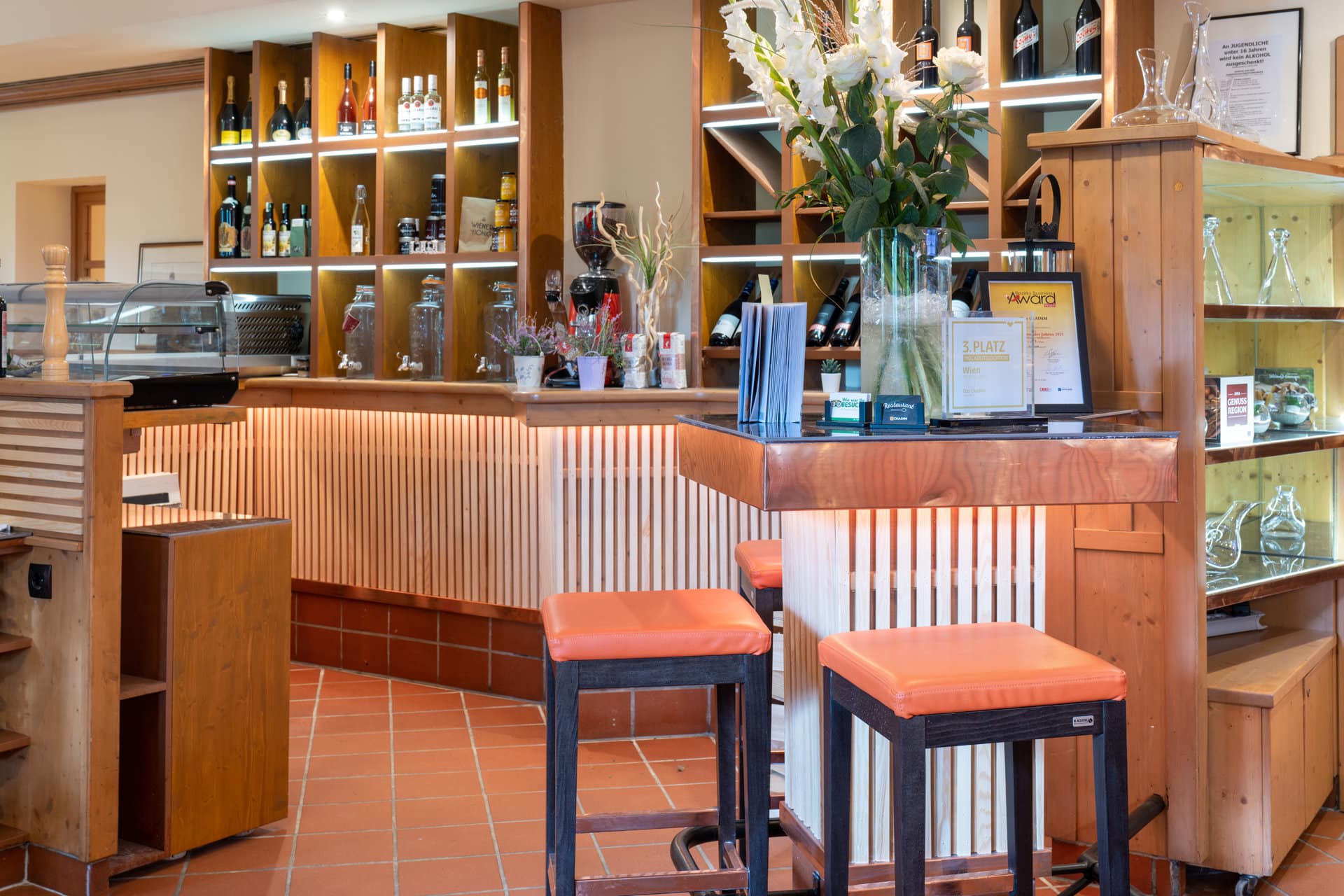 20220908_9332 Gemütlicher Barbereich im Restaurant Das Chadim mit Holzregalen, Getränken, Blumenarrangement und orangefarbenen Barhockern, ideal für Veranstaltungen und Feiern.
