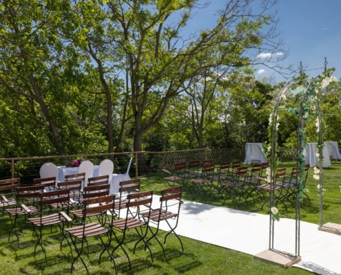 © Adrian Almasan | www.adrianalmasan.com Hochzeitszeremonie im Freien mit Stühlen und einem geschmückten Bogen, umgeben von Bäumen im Chadim-Garten.