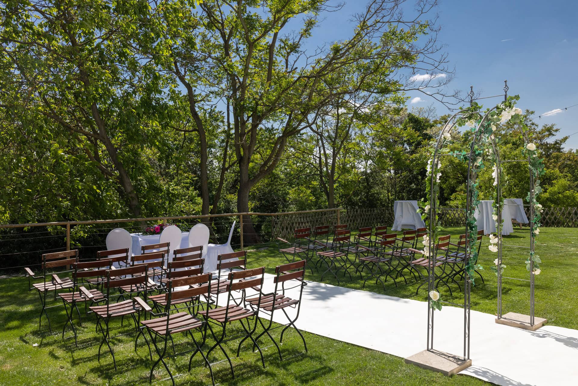 © Adrian Almasan | www.adrianalmasan.com Hochzeitszeremonie im Freien mit Holzstühlen, einem weißen Teppich und einer mit Blumen geschmückten Bogenarchitektur, umgeben von grünen Bäumen im Gastgarten von Das Chadim.