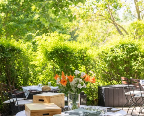 © Adrian Almasan | www.adrianalmasan.com Blühende Blumenarrangements auf einem Tisch im Gastgarten von Das Chadim, umgeben von grünen Pflanzen und schattigen Sitzgelegenheiten, ideal für Events und Feiern.