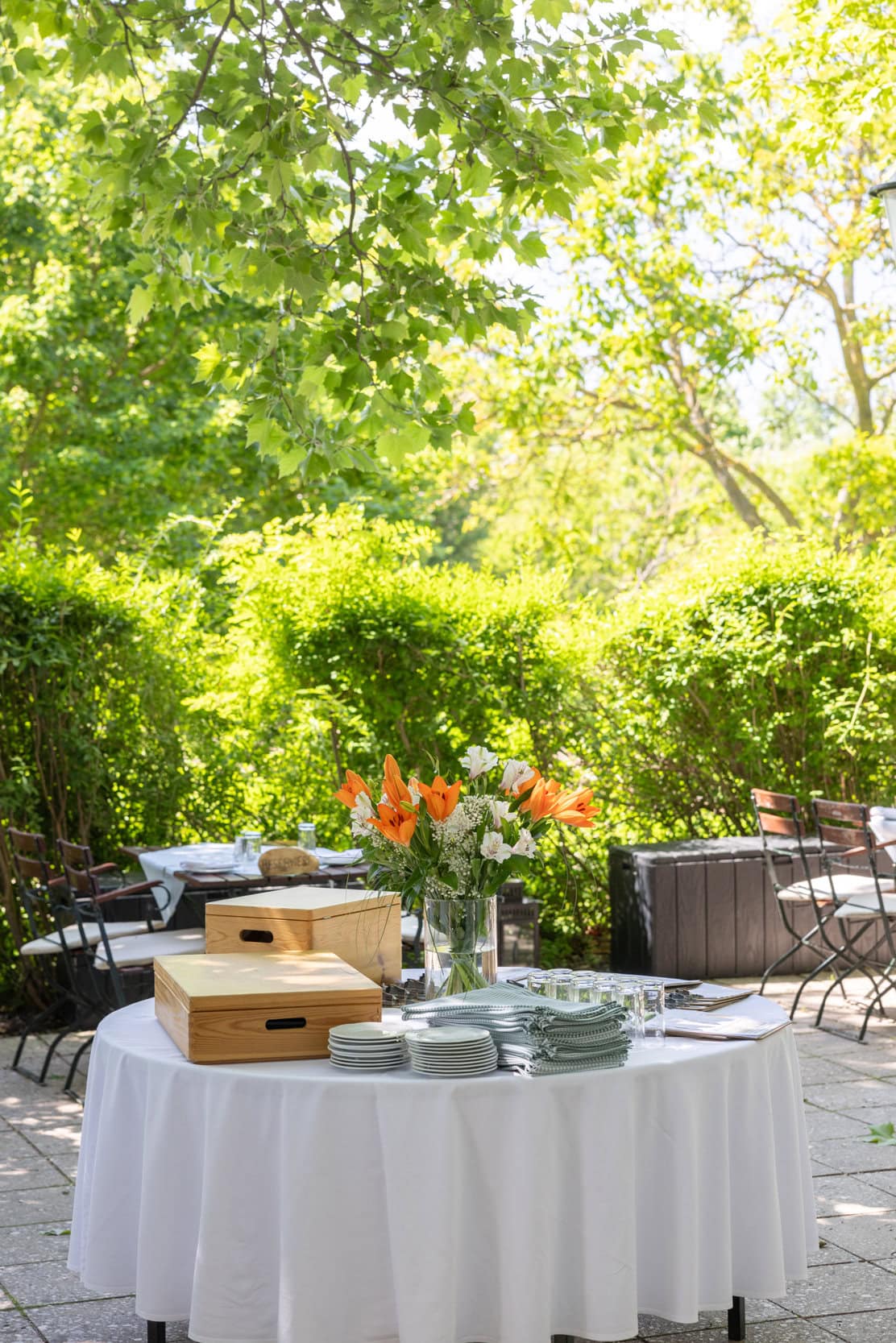 © Adrian Almasan | www.adrianalmasan.com Gedeckter Tisch im Gastgarten von Das Chadim mit frischen Blumen, Geschirr und Holzboxen, umgeben von grüner Natur und schattigen Bäumen.