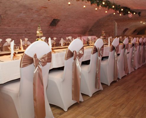 Gewölbe_dekoriert Elegant dekorierter Festsaal mit langen Tischen, weißen Stuhlhussen und goldenen Schleifen, ideal für Veranstaltungen im historischen Gewölbe von Das Chadim.