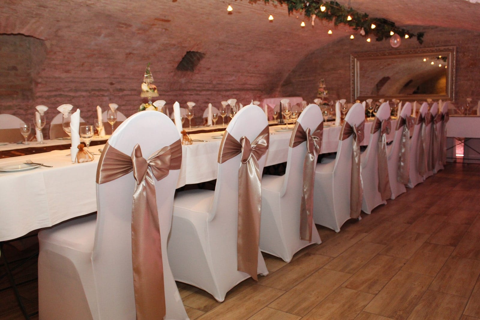 Gewölbe_dekoriert Elegant dekorierter Bankettraum im historischen Gewölbe mit festlich gedecktem Tisch, weißen Stühlen mit goldenen Schleifen und sanfter Beleuchtung.