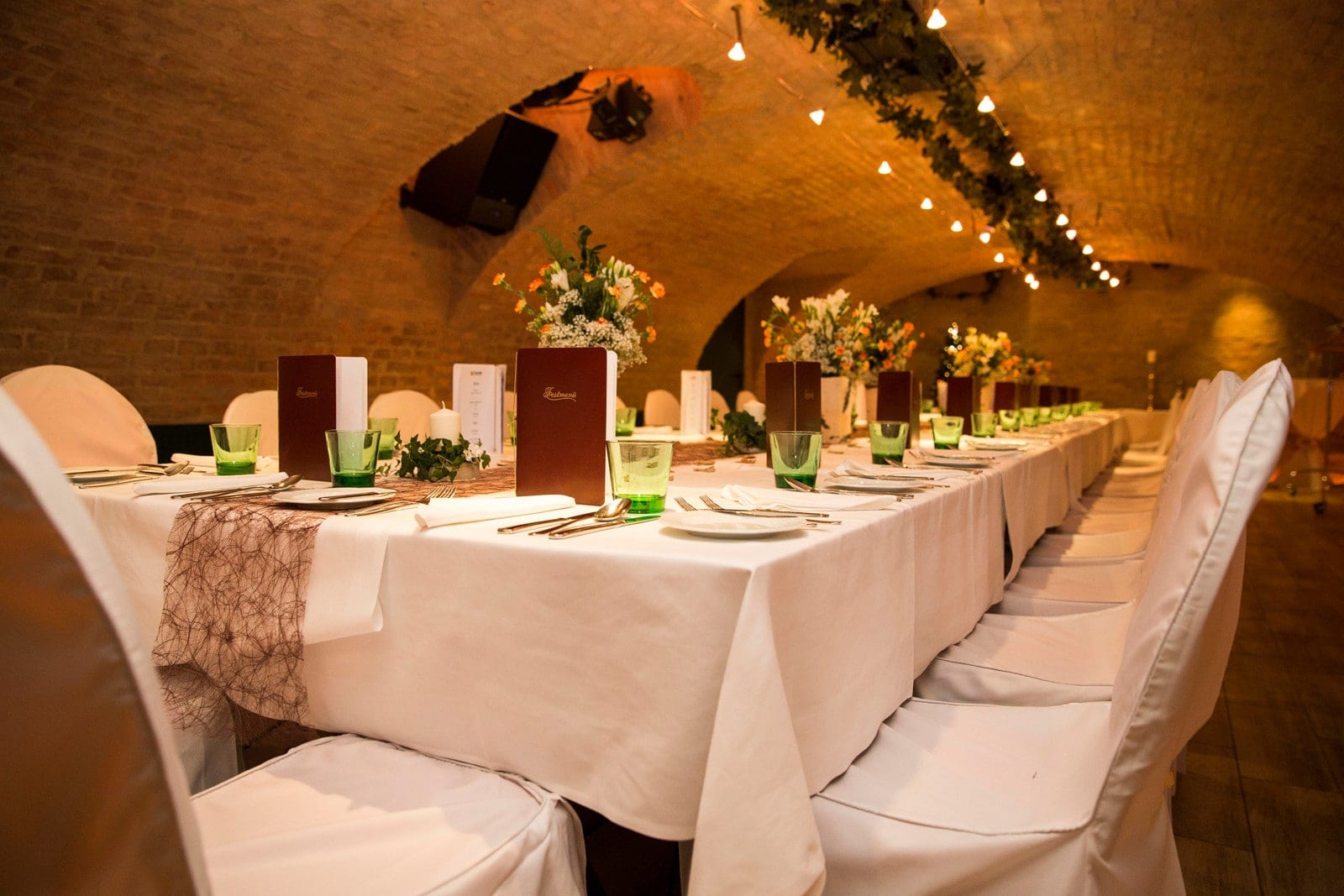 Tafel_Gewölbe_Hochzeit_vh Elegant Bankettraum im historischen Gewölbe von Das Chadim, festlich gedeckte Tische mit Blumenarrangements, Menükarten und grünem Glas, ideal für Hochzeiten und Feiern.