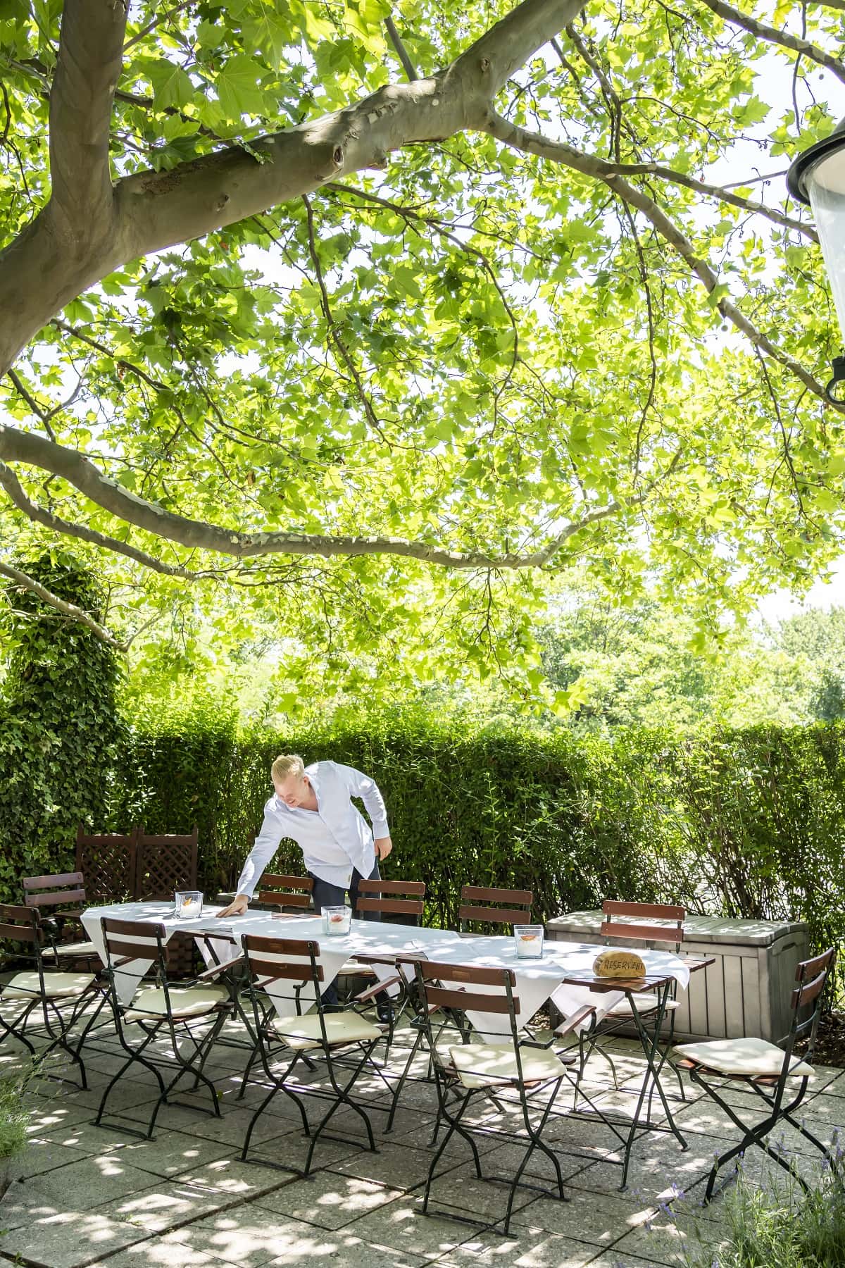 herbst-6200 Gastgarten von Das Chadim mit gedecktem Tisch unter schattigen Bäumen, Mitarbeiter bereitet die Tafel für Gäste vor, idyllische Umgebung mit Blick ins Grüne.