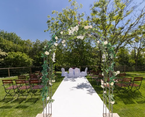 startseite_slider_heiraten Hochzeitszeremonie im Freien mit einem Blumenbogen, weißen Stühlen und einer festlichen Atmosphäre im Garten von Das Chadim.